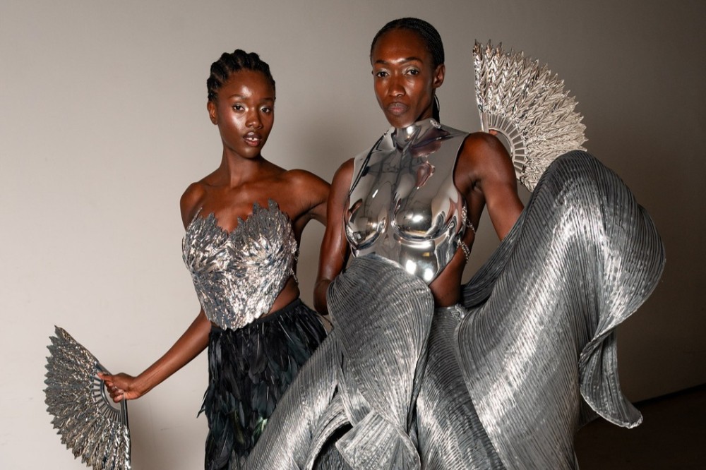 Duas modelos negras posam com roupas futuristas prateadas. A da esquerda usa um corpete texturizado e saia de penas, segurando um leque. A da direita veste um top metálico e calça plissada, com um leque maior atrás