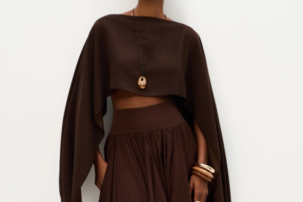 Mulher de pele escura vestindo um conjunto marrom escuro, composto por um cropped de mangas longas e amplas, e uma saia longa com cintura franzida. Ela usa um colar com pingente oval de madeira e pulseiras de madeira no braço direito, com as mãos nos bolsos da saia