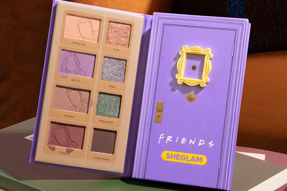Paleta de sombras Sheglam Friends aberta, com um lado roxo imitando a porta do apartamento da série, com moldura amarela e a palavra FRIENDS. O outro lado tem oito sombras em tons de nude, roxo, verde e marrom, algumas com desenhos dos personagens e nomes como Turkey Hat e Apt 20