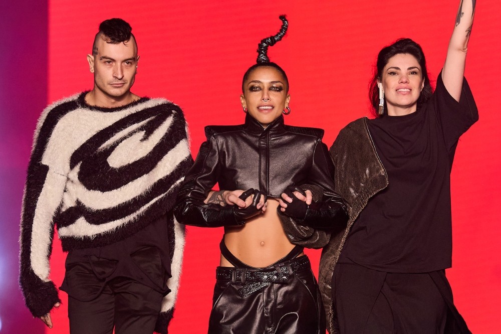 Três pessoas posam sorrindo em um palco vermelho. À esquerda, um homem de moicano e suéter preto e branco. No centro, uma mulher de top e calça de couro preta, com cabelo em espiral. À direita, uma mulher de camiseta preta e jaqueta marrom no ombro, braço esquerdo levantado