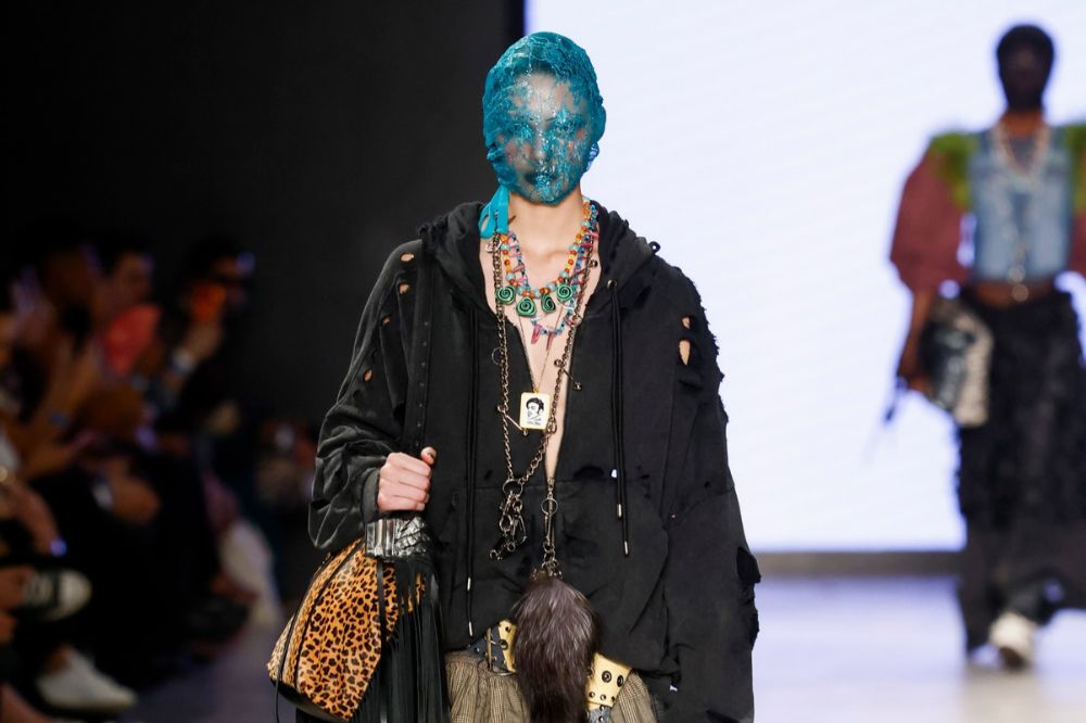 Modelo desfila com rosto coberto por véu azul-turquesa, usando moletom preto rasgado, colares coloridos e bolsa de estampa animal print com franjas pretas, em passarela de moda