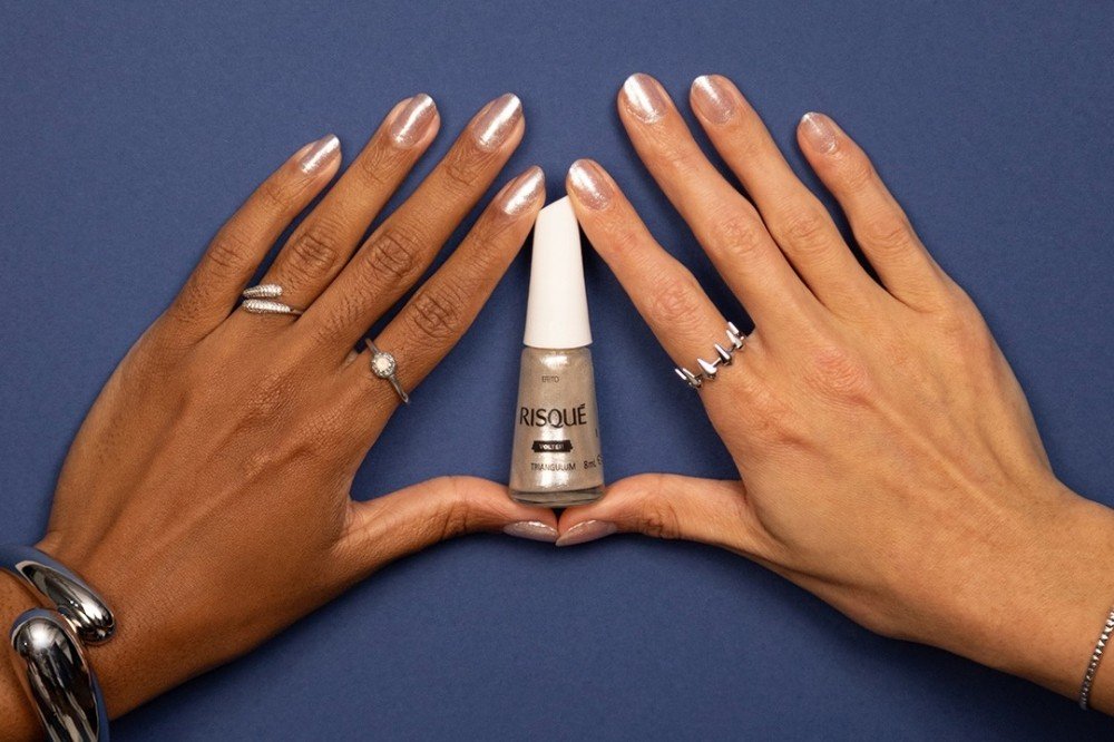 Duas mãos femininas, uma de pele morena e outra clara, com unhas pintadas de esmalte perolado, seguram um frasco de esmalte Risqué na cor As Mil Purpurinas sobre um fundo azul escuro. A mão morena usa anéis prateados e uma pulseira grossa, enquanto a mão clara exibe um anel de spikes e uma pulseira fina