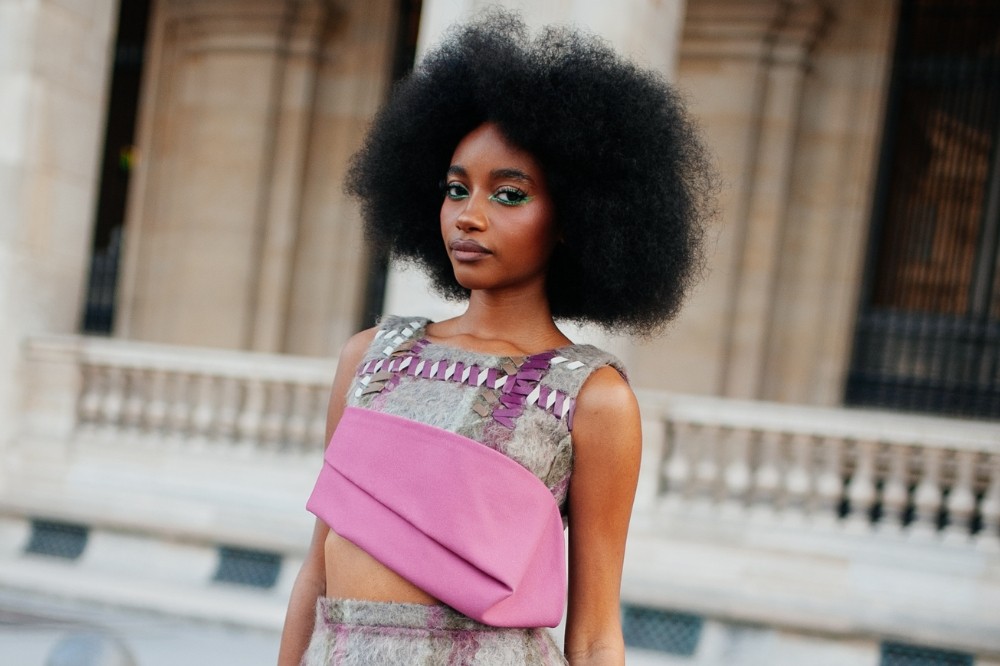 Mulher negra com cabelo afro volumoso, maquiagem verde nos olhos, vestindo um top cropped cinza com detalhes roxos e uma faixa rosa na cintura, em frente a um prédio claro com colunas