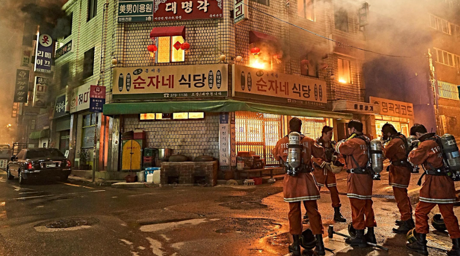 Bombeiros com uniformes laranja e tanques de oxigênio nas costas se reúnem em uma rua escura, observando um prédio de tijolos com chamas e fumaça saindo das janelas do segundo andar. Placas em coreano adornam a fachada, e um carro escuro está estacionado à esquerda.