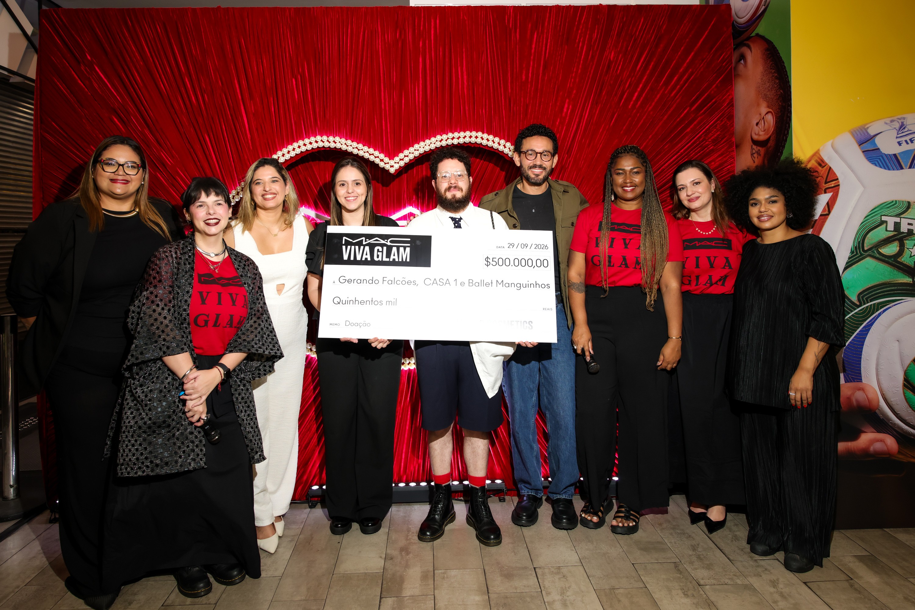 Dez pessoas sorridentes, incluindo homens e mulheres, posam em frente a um fundo vermelho com um coração. O grupo segura um cheque gigante da MAC VIVA GLAM de R$ 500.000,00 para Gerando Falcões e CASA 1º Ballet Manguinhos. Algumas mulheres vestem camisetas vermelhas com a inscrição VIVA GLAM