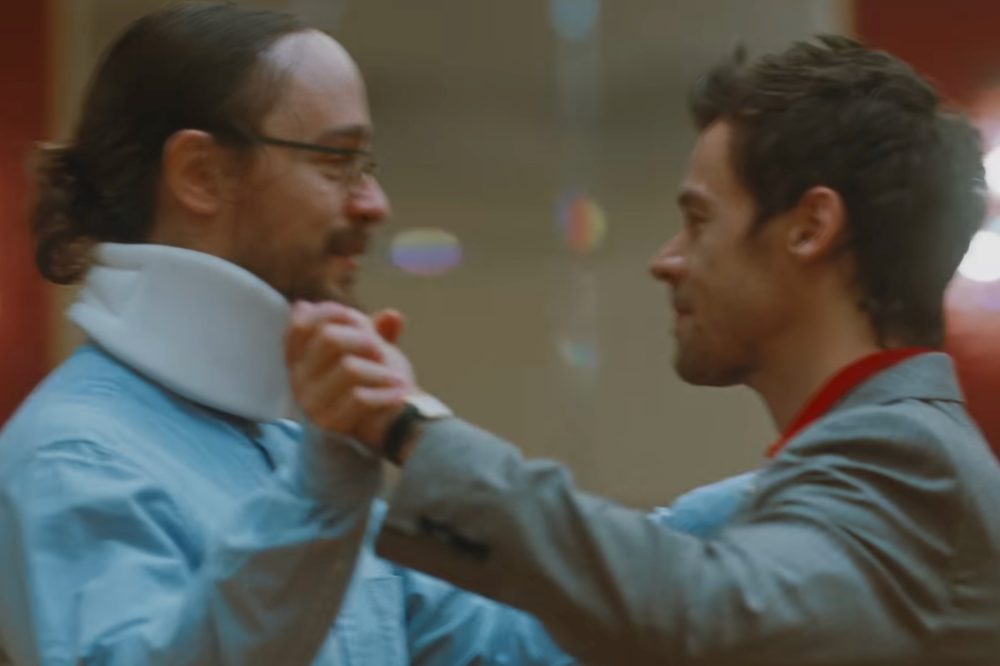Dois homens sorrindo, um de perfil com colar cervical branco e camisa azul, e outro de terno cinza com camisa vermelha, dançando e segurando as mãos