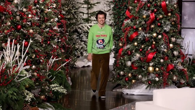 Harry Styles sorrindo, caminhando em um palco decorado com várias árvores de Natal iluminadas, cheias de bolas vermelhas, douradas e prateadas, fitas vermelhas e folhagens brancas. Ele veste um suéter verde-limão com a frase Stay away from toxic people e calças marrons.