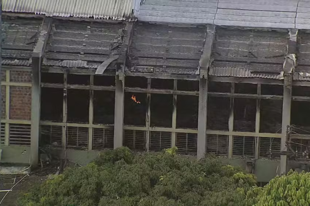 Fachada de um prédio industrial com telhado danificado e janelas escuras, mostrando sinais de incêndio. Chamas alaranjadas e pequenas são visíveis em uma das janelas do andar superior. Abaixo, arbustos verdes cobrem parte da base da construção