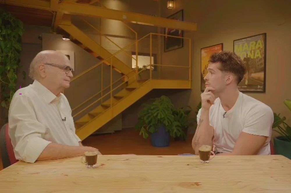 Dois homens sentados em uma mesa de madeira clara, com xícaras de café. O mais velho, de camisa branca e óculos, olha para o jovem, de camiseta branca, que apoia o queixo na mão, pensativo. Ao fundo, uma escada amarela e plantas.