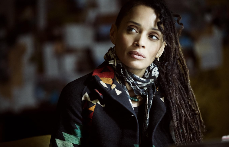 Mulher de pele clara e dreadlocks escuros, com expressão pensativa, usando um casaco preto com estampa geométrica colorida e um lenço estampado em preto e branco, olhando para o lado direito