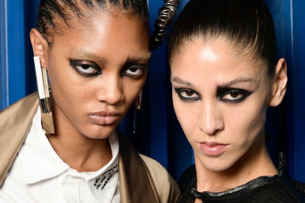 Duas modelos com maquiagem dramática nos olhos, uma com tranças e brincos longos, a outra com cabelo puxado para trás, ambas com expressão séria, em fundo azul