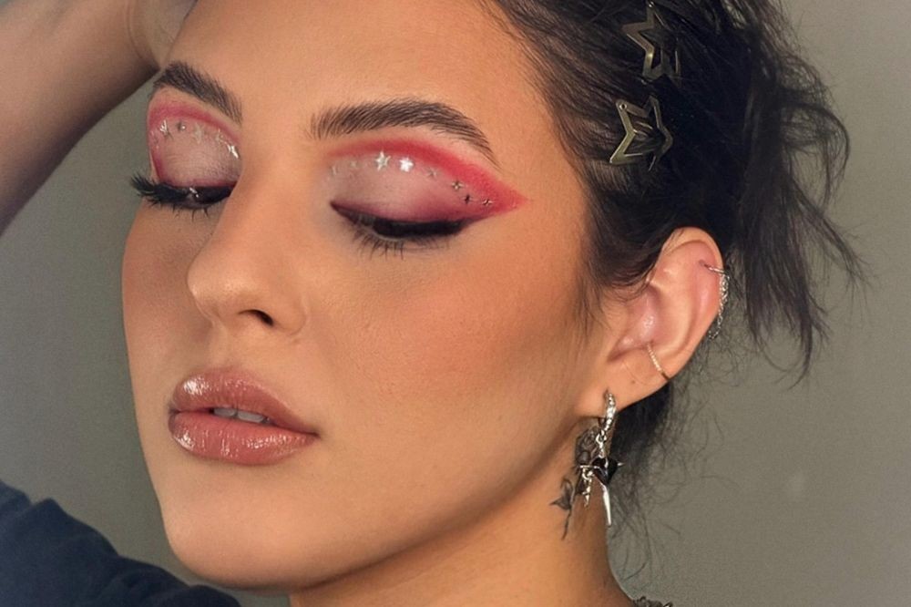 Close-up de uma mulher com maquiagem artística nos olhos, pálpebras fechadas, sombra rosa e branca com glitter de estrelas, delineado gráfico. Ela tem lábios com gloss, brincos prateados e presilhas de estrela no cabelo escuro preso.