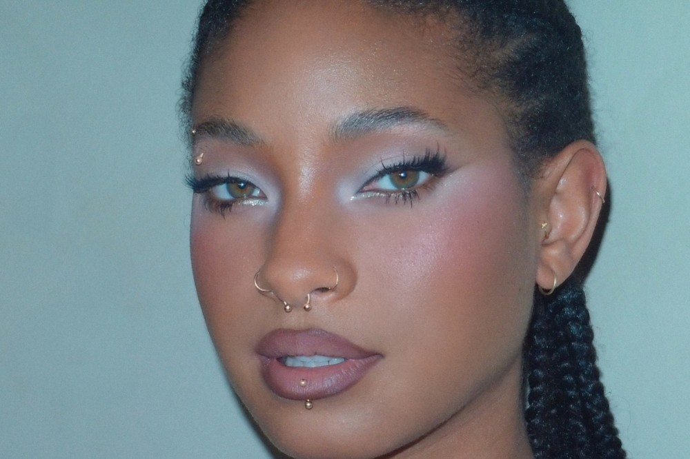 Rosto de uma mulher jovem com pele morena, olhos castanhos esverdeados, maquiagem rosa e branca, piercing no nariz e lábio inferior, e tranças finas no cabelo