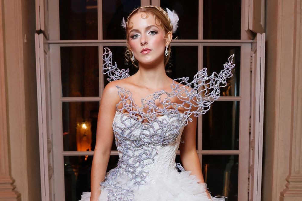 Mulher loira com penteado elaborado e penas brancas, usando um vestido branco com detalhes prateados brilhantes, posa séria em frente a uma janela com moldura branca à noite