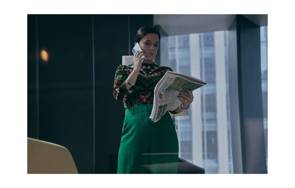 Mulher de cabelos escuros, blusa estampada e calça verde, fala ao telefone enquanto lê um jornal, em um escritório moderno com janelas grandes