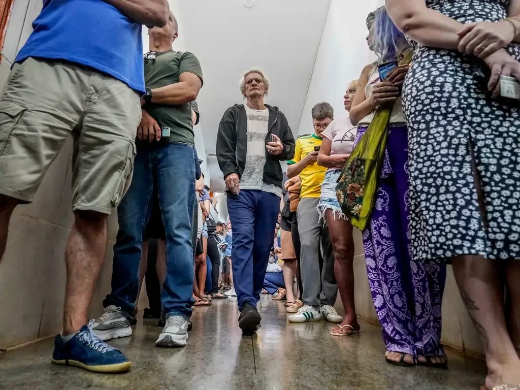 Pessoas de diversas idades e etnias em uma fila, algumas olhando para cima, outras para seus celulares. Um homem de cabelos grisalhos e camisa listrada caminha em direção ao observador. O chão é de cimento polido e as paredes brancas, sugerindo um ambiente interno. A imagem é cortada na altura do peito das pessoas, focando nas pernas e pés