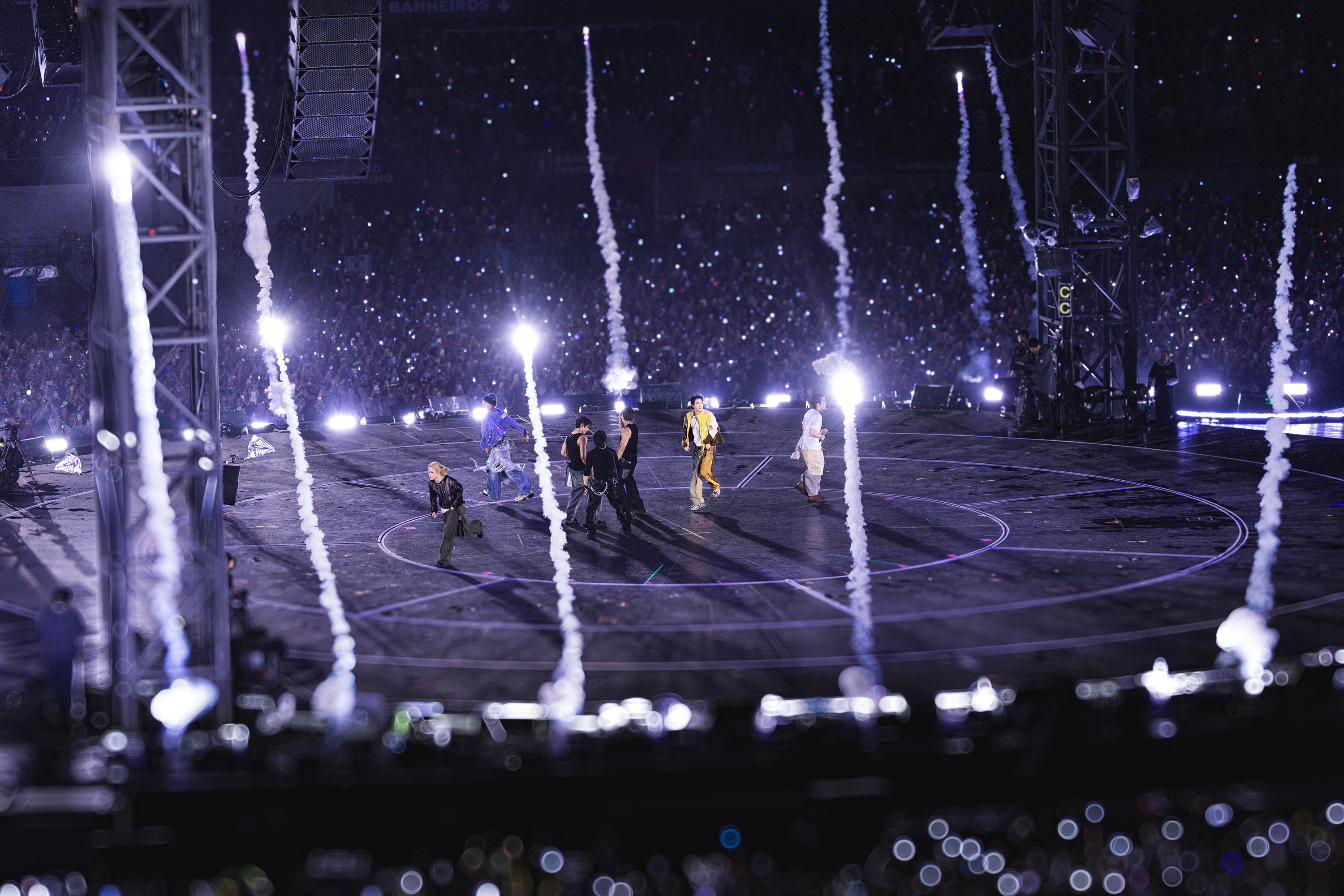 Sete artistas performam em palco escuro com círculos roxos, enquanto fogos de artifício verticais brancos sobem. A plateia ao fundo ilumina o estádio com luzes de celular
