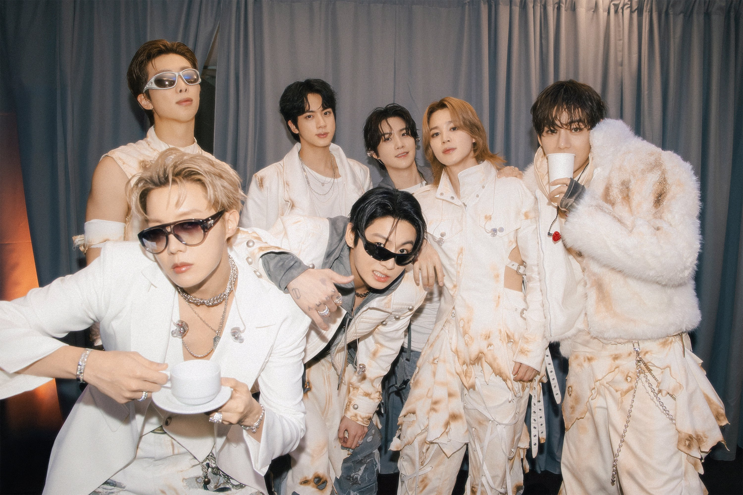 Sete homens jovens, membros do grupo BTS, posam com roupas brancas e bege de estilo futurista e desgastado. Alguns usam óculos escuros, outros seguram xícaras. O fundo é cinza com cortinas.