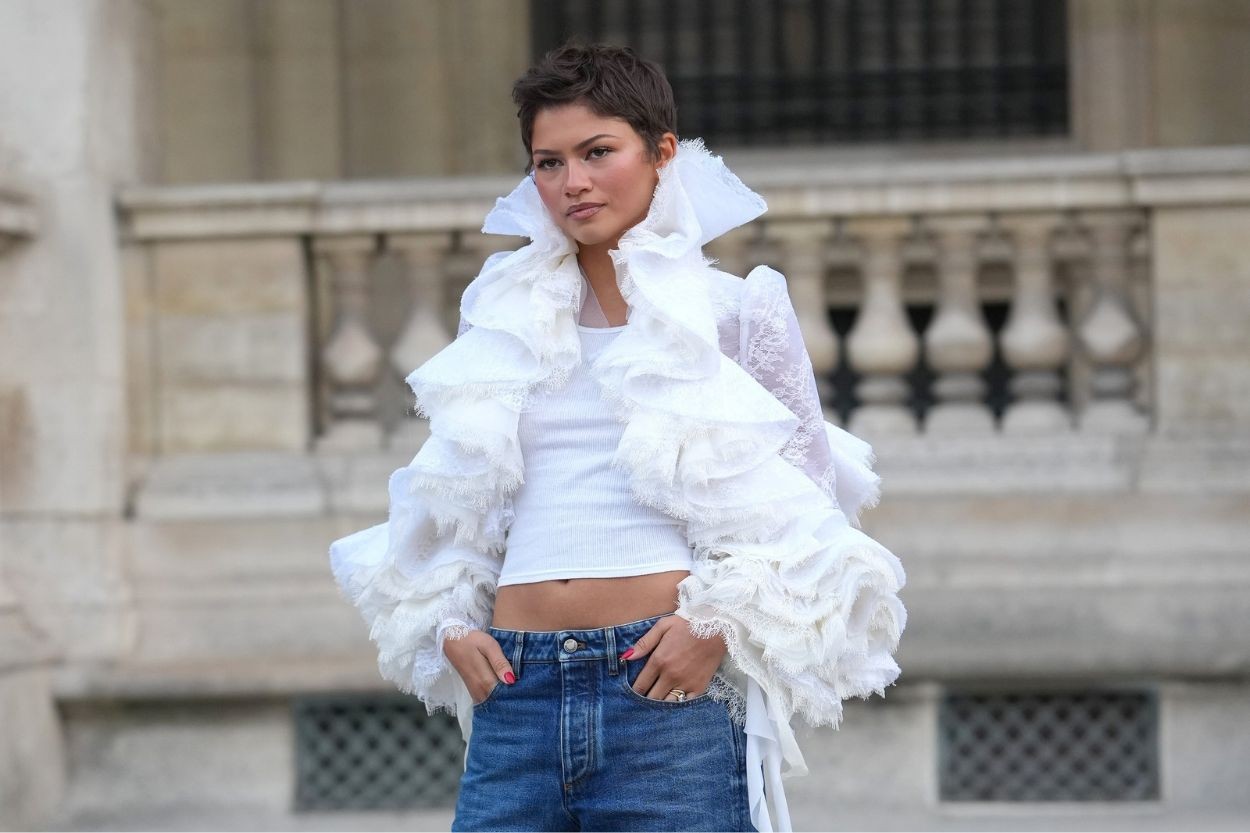 Zendaya com cabelo curto e escuro, vestindo um top branco justo e um casaco branco volumoso com babados e mangas de renda, calça jeans e unhas pintadas de rosa, posando em frente a um edifício de pedra com balaustrada