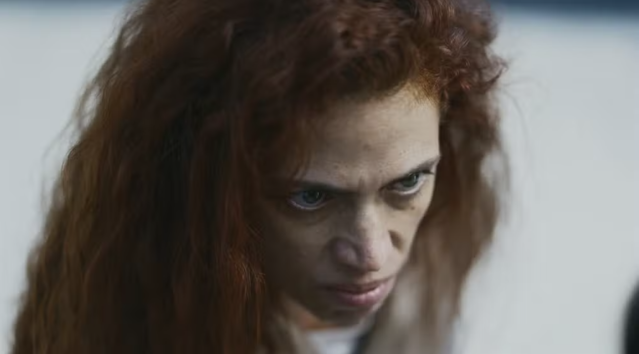 Mulher de cabelos ruivos e encaracolados, com expressão séria e olhar fixo para baixo, sobrancelhas franzidas e olhos claros, em um ambiente com fundo claro e desfocado